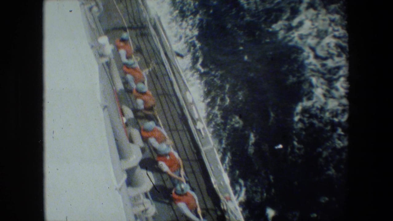 High-angle shipboard view of sailors in orange life vests lined along a deck rail while handling a rope or line beside foaming ocean water.