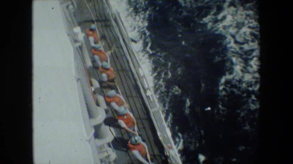 High-angle shipboard view of sailors in orange life vests lined along a deck rail while handling a rope or line beside foaming ocean water.