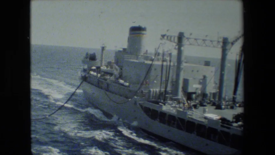 Soft 8mm frame of a large vessel at sea, showing deck structures, railings, a smokestack-like structure, rigging, and foaming wake behind the ship.