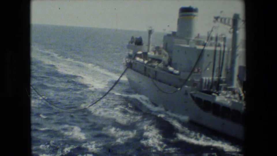 Archival 8mm view of a large ship underway, with ocean wake, deck equipment, railings, rigging, and lines visible along the vessel's side.