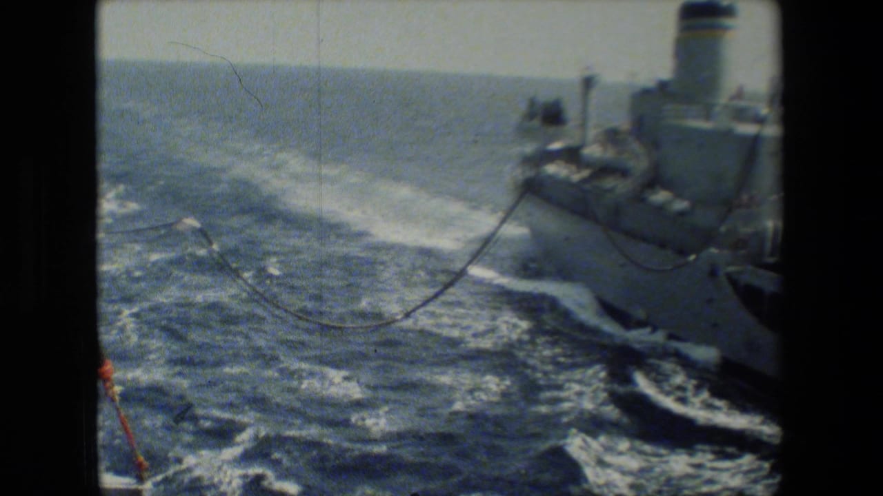 A deck-level 8mm view shows a naval vessel alongside another ship at sea, with rigging or transfer line visible over choppy water.