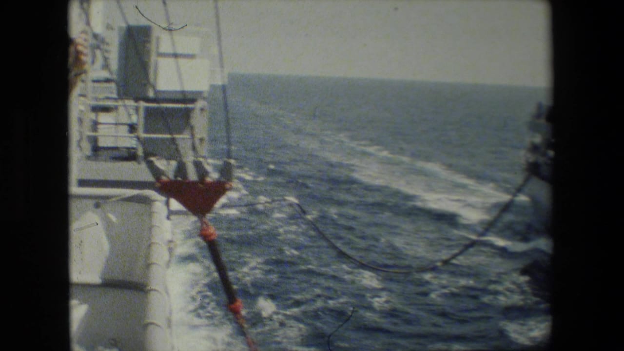 Shipboard deck view over open water with a suspended cable or hose and wake alongside.