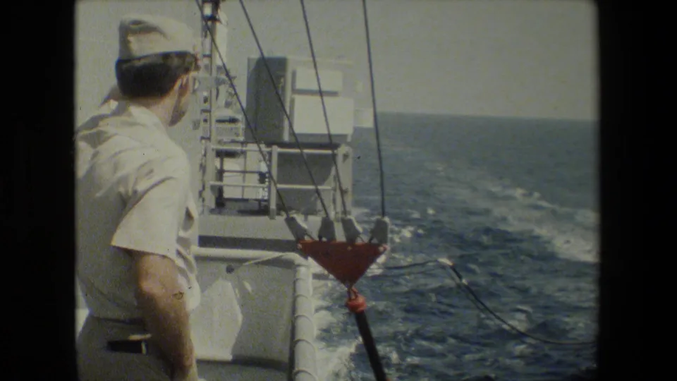 A sailor in a white uniform and cap stands on deck looking toward the transfer rigging and ocean during the naval operation.