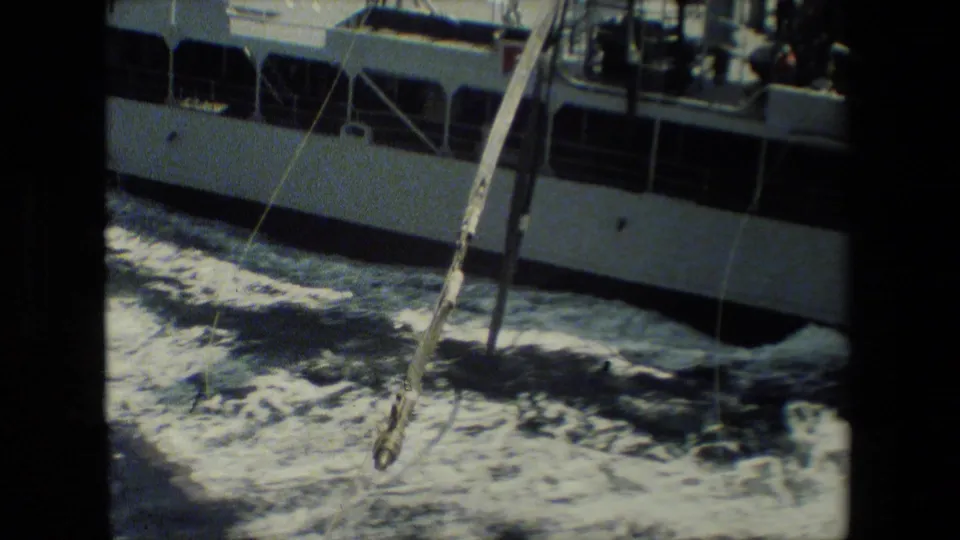 A neighboring naval ship's hull passes close by as a line or hose hangs over foamy water between the vessels.