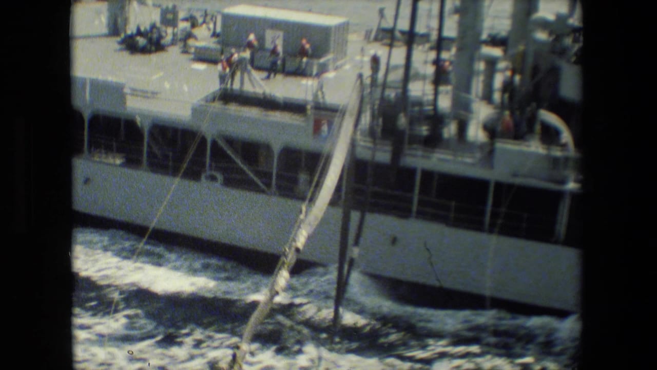 A vessel seen from close range with crew members on deck near railings and rigging, with white hull sections and churning seawater below.