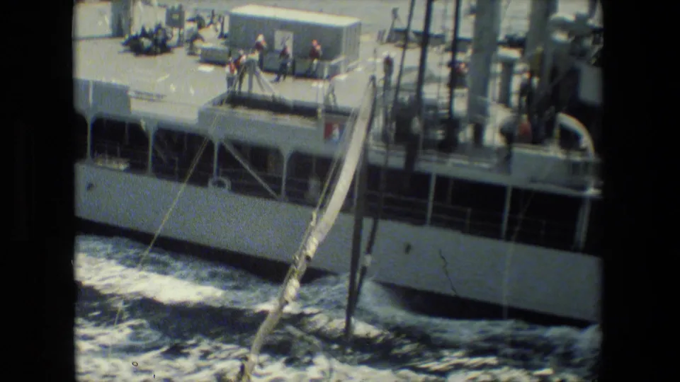 A vessel seen from close range with crew members on deck near railings and rigging, with white hull sections and churning seawater below.