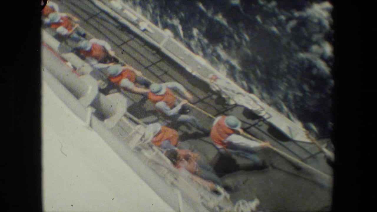 Crew members in orange flotation vests and grey uniforms appear to pull a heavy line together along the sloped deck of a vessel, with dark water beside the hull.