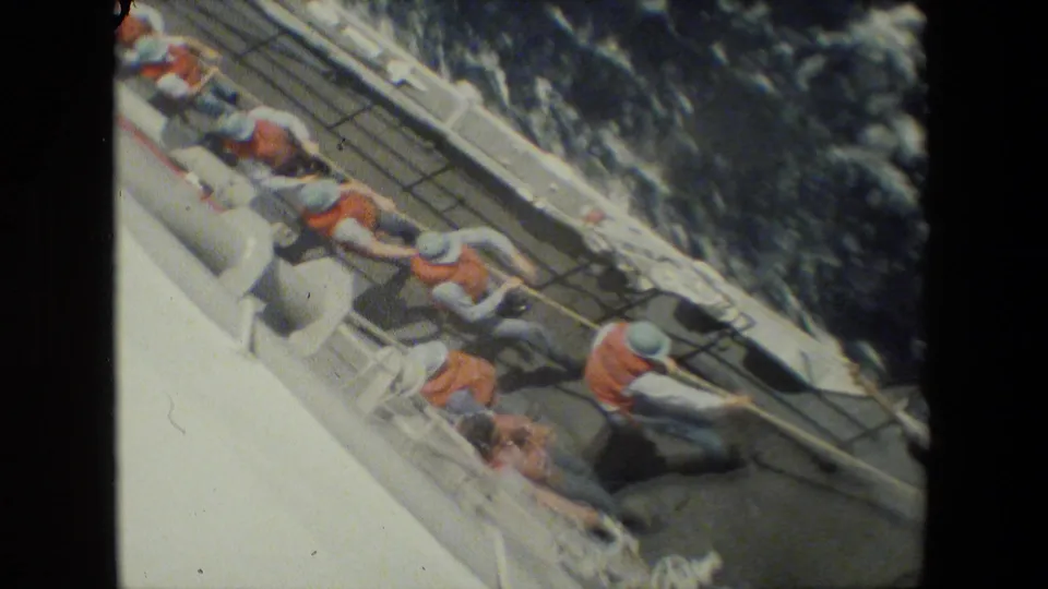 Crew members in orange flotation vests and grey uniforms appear to pull a heavy line together along the sloped deck of a vessel, with dark water beside the hull.