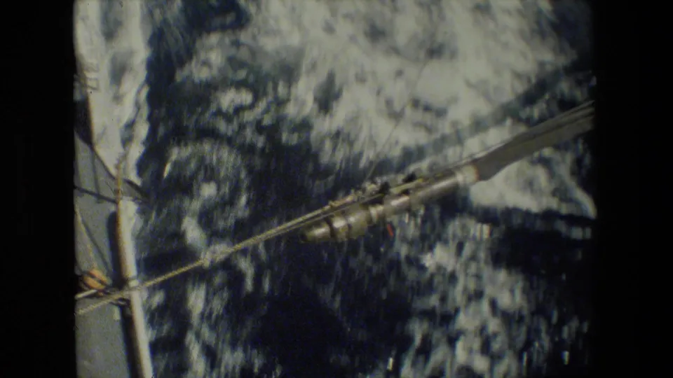 A close handheld view shows shipboard lines, railings, and deck hardware passing over foaming blue water beside the vessel.