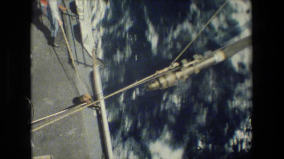 Diagonal ship railings and taut lines frame a close view of rough ocean water along the side of the vessel, with no clear individual faces visible in this frame.