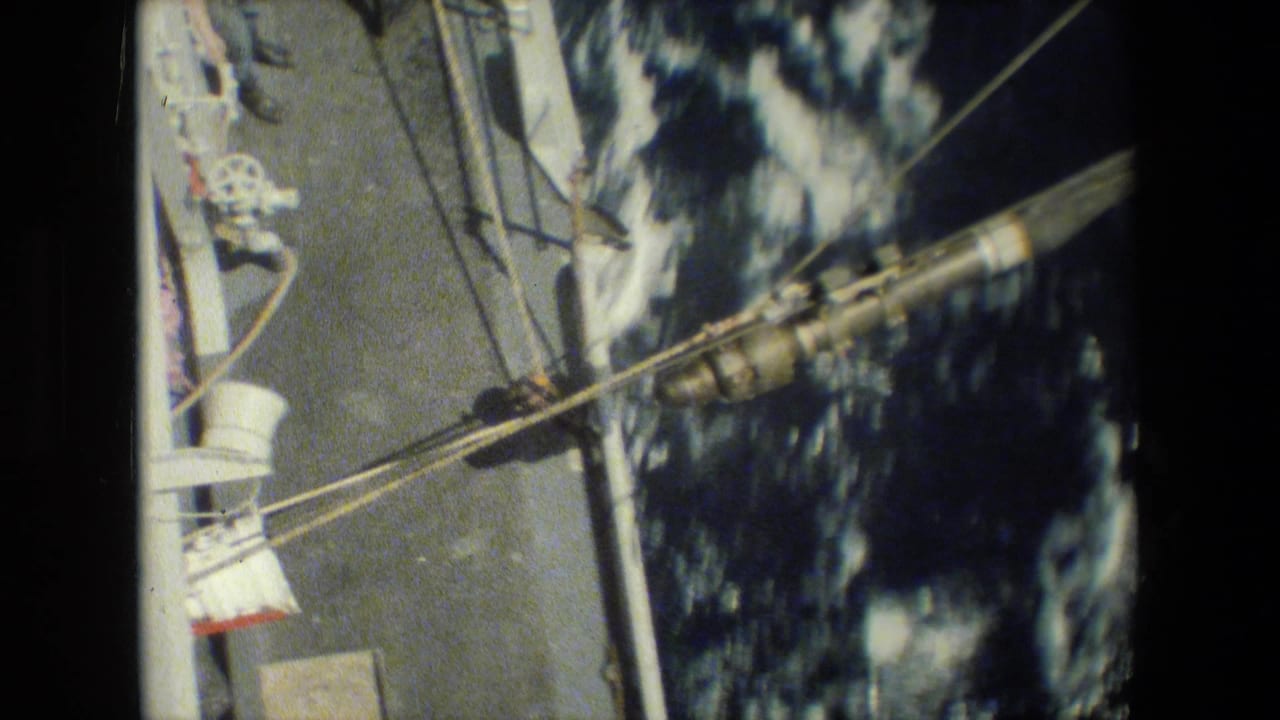 Overhead 8mm frame showing deck rigging, cables, a white structural post, mechanical fittings, and dark choppy water alongside the ship.