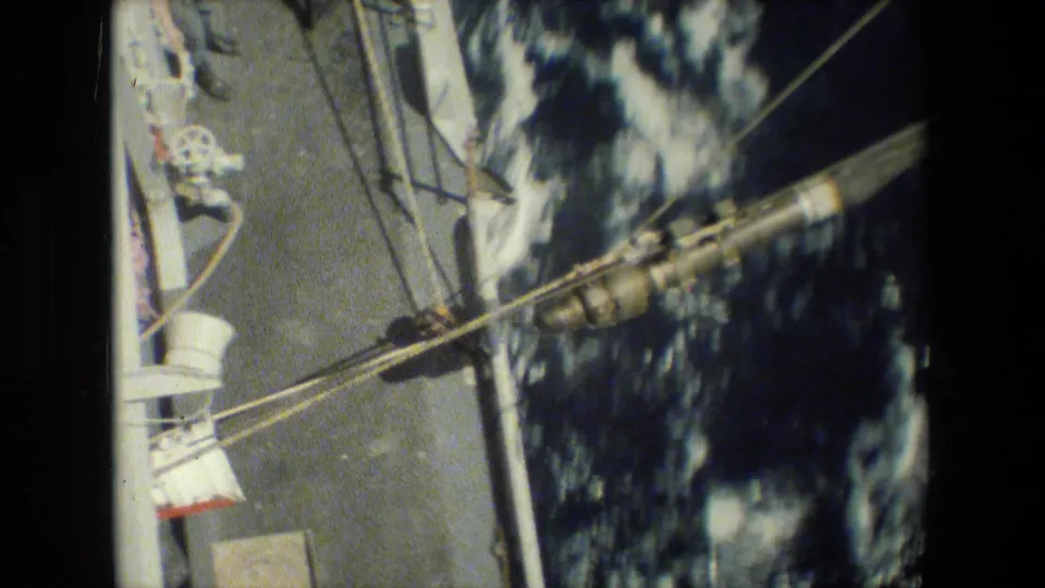Overhead 8mm frame showing deck rigging, cables, a white structural post, mechanical fittings, and dark choppy water alongside the ship.