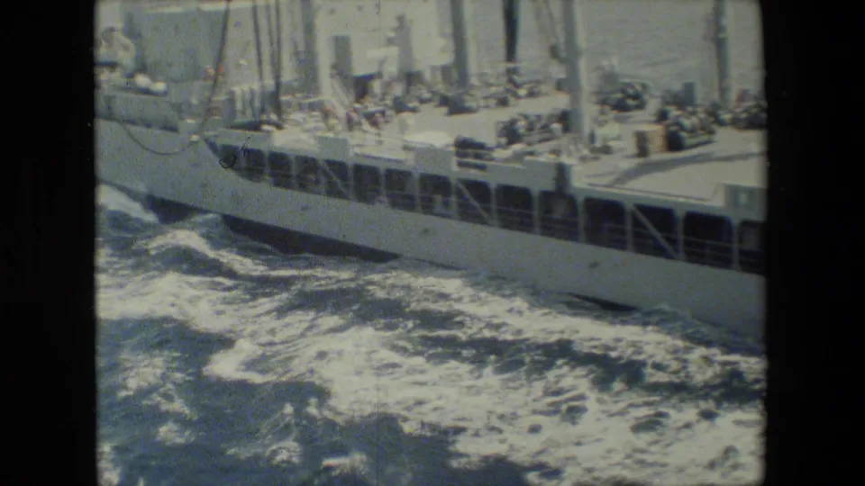 Color 8mm frame showing the side of a ship with railings, deck structures, and foamy choppy water along the hull.