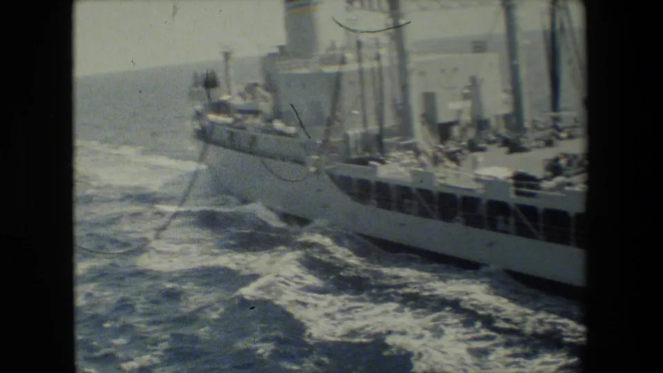 Side view of a vessel section with railings, deck equipment, vertical posts, and rough blue water breaking alongside the hull.
