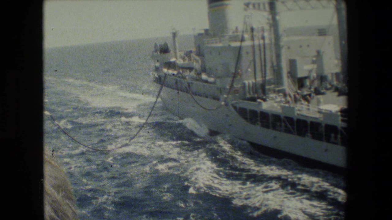 A gray naval-looking vessel travels alongside another ship on open water, with wake, deck structures, and possible connecting lines visible in grainy 8mm footage.