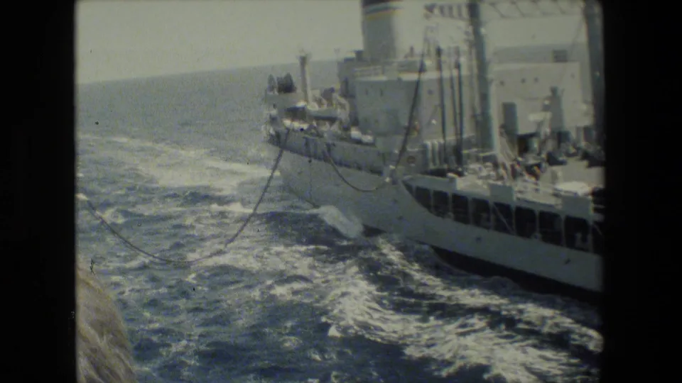 A gray naval-looking vessel travels alongside another ship on open water, with wake, deck structures, and possible connecting lines visible in grainy 8mm footage.