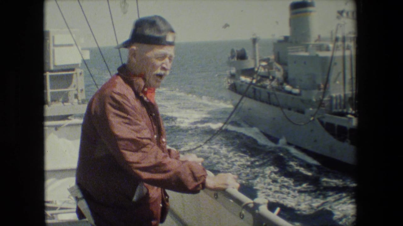 An older man wearing a brown jacket and backward cap stands at a ship railing while a gray vessel and foamy wake pass behind him at sea.