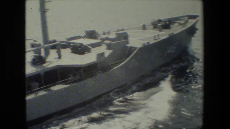 A gray vessel is seen from above and alongside, showing its bow, deck hardware, hull side, and foamy water in a handheld 8mm maritime view.