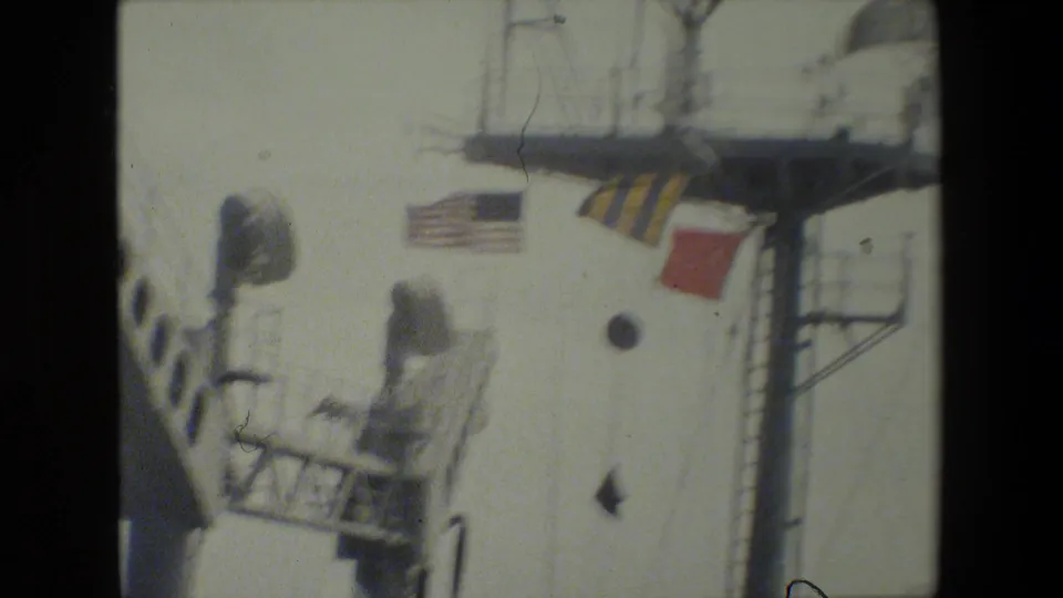 1981 8mm home-movie frame showing part of a naval ship superstructure with a U.S. flag, colored signal flags, railings, ladders, and bright sky.