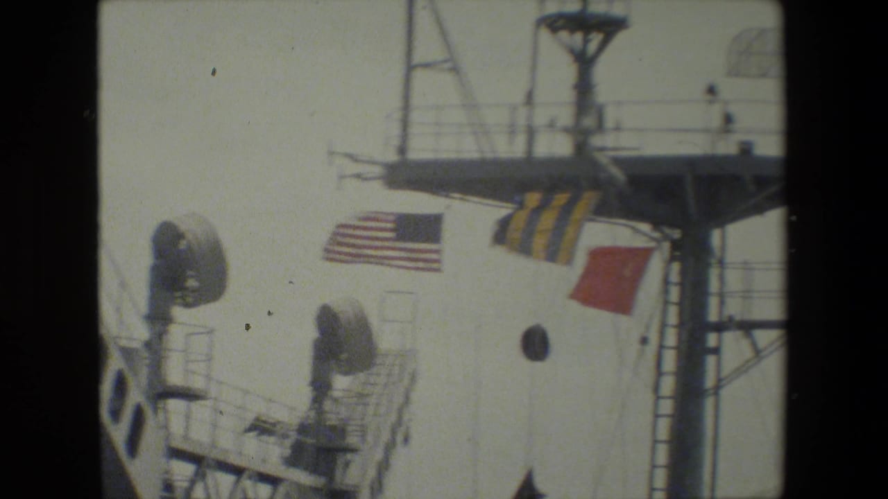 Close shipboard view with a U.S. flag, signal flags, mast platform, circular porthole, ladder, and railings visible in soft 8mm focus.