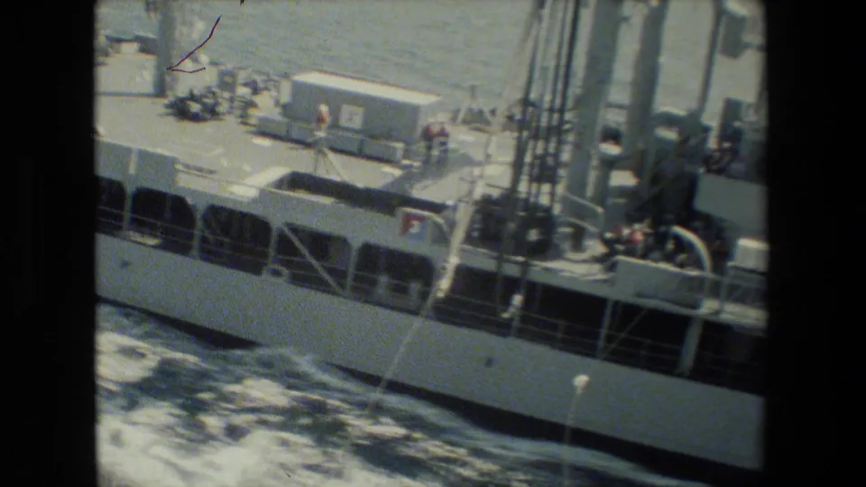 Home-movie frame looking down across a ship deck with crew figures, deck equipment, railings, mast supports, and ocean water visible along the hull.