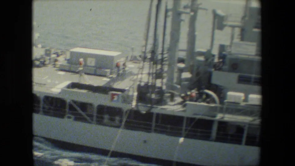 Oblique 8mm view across a naval ship deck showing small crew figures, railings, vertical mast supports, deck equipment, hull openings, and churning water.