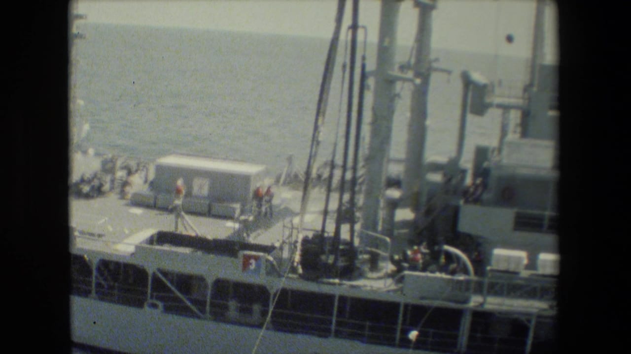 8mm frame of a gray military vessel seen from above, with crew on deck near railings, mast structures, and deck equipment at sea.