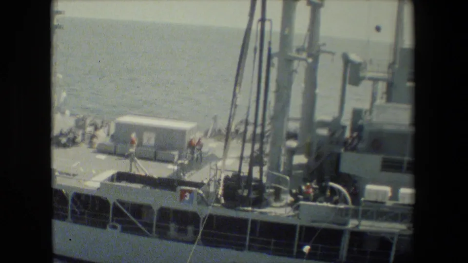 8mm frame of a gray military vessel seen from above, with crew on deck near railings, mast structures, and deck equipment at sea.