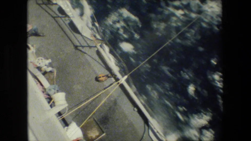 Oblique 8mm frame of the vessel's side and deck hardware, with taut lines crossing toward churning water alongside the hull.