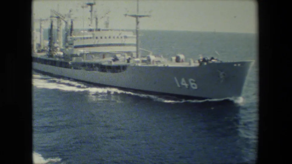 A large white naval vessel with hull number 146 passes across blue open water, with deck equipment, mast structures, and a foaming wake visible in grainy 8mm footage.