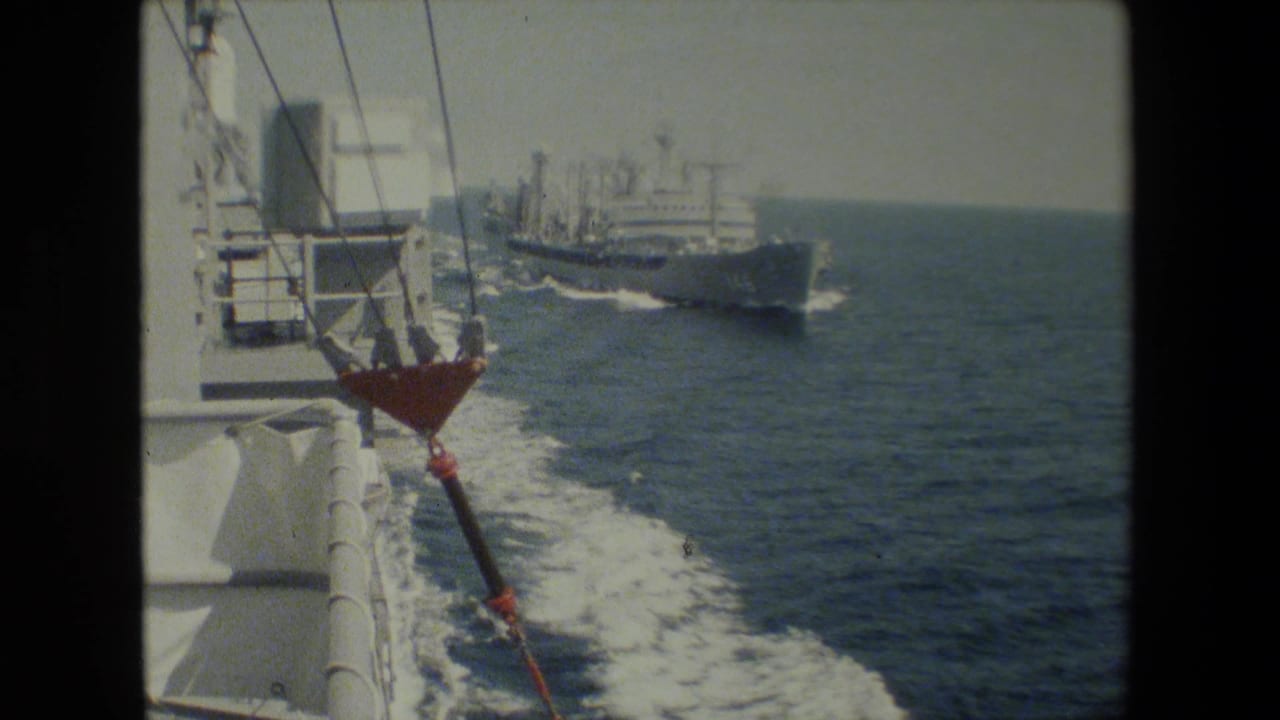 Naval ship seen from another vessel with deck railing and rigging in foreground.
