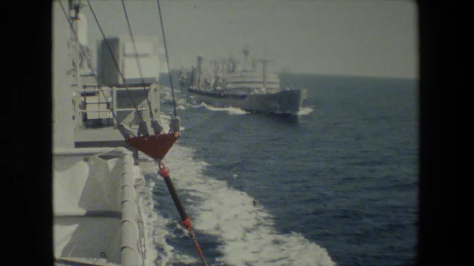 A gray naval ship travels off the side of the filming vessel, with railings, rigging, a red deck fitting, open sea, and the ship's wake visible in the frame.