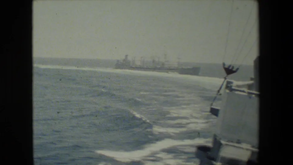 A naval ship appears in the distance beyond a wide curving wake, with part of the filming vessel's side structure and rigging visible along the right edge.