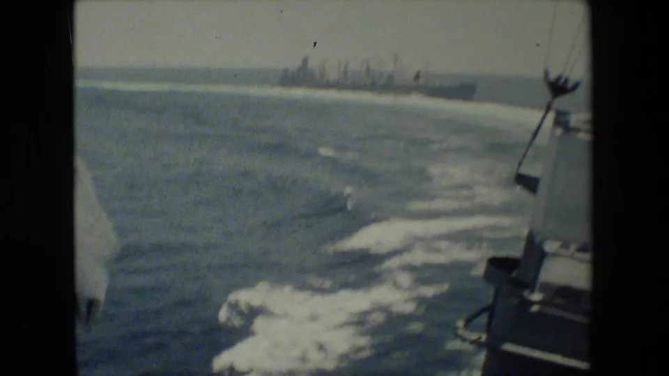 A gray naval vessel sits far across the water while the filming vessel's wake curves through the foreground and shipboard fixtures appear at the right edge.