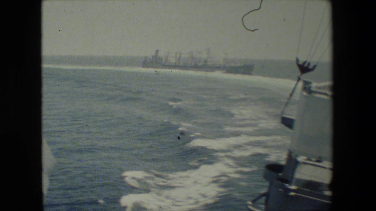 Faded 8mm frame showing open ocean, foamy ship wake, partial ship structure at right, and distant naval vessels on the horizon.