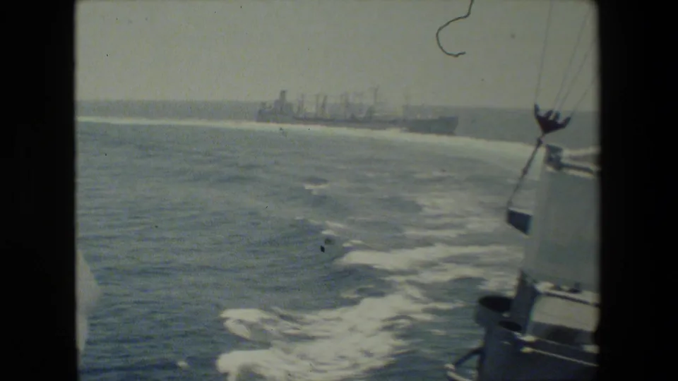 Faded 8mm frame showing open ocean, foamy ship wake, partial ship structure at right, and distant naval vessels on the horizon.