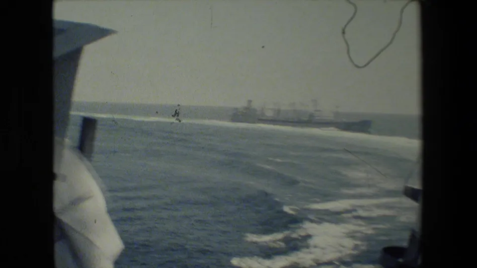 Shipboard view across calm ocean water and wake toward distant gray naval vessels, with part of the filming vessel visible at the right edge.