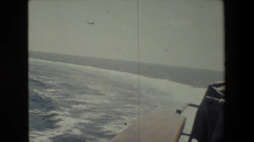 Faded 8mm shipboard frame looking down along foamy wake, with a partial deck edge and a low distant horizon or coastline visible.