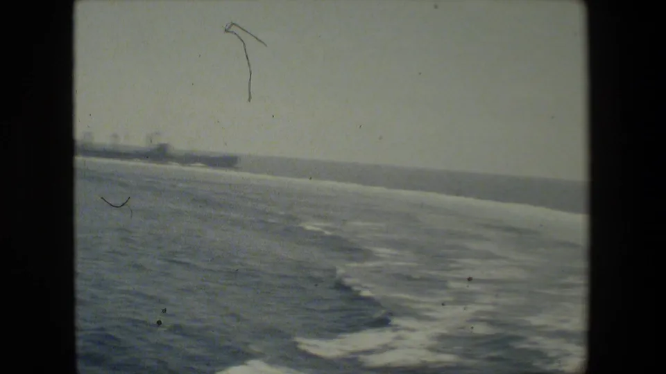 Soft, color-faded 8mm frame of ocean wake curving across the water, with faint distant ship silhouettes along the horizon.