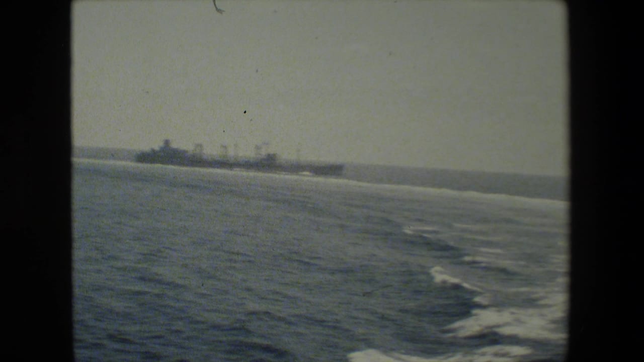 A distant naval vessel travels across open water, seen from another ship or elevated vantage point in grainy 8mm footage.
