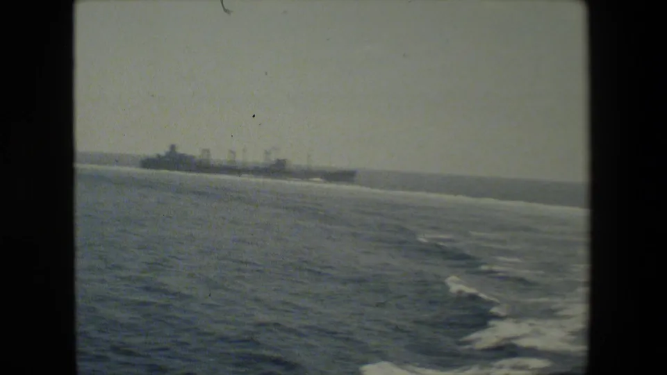 A distant naval vessel travels across open water, seen from another ship or elevated vantage point in grainy 8mm footage.