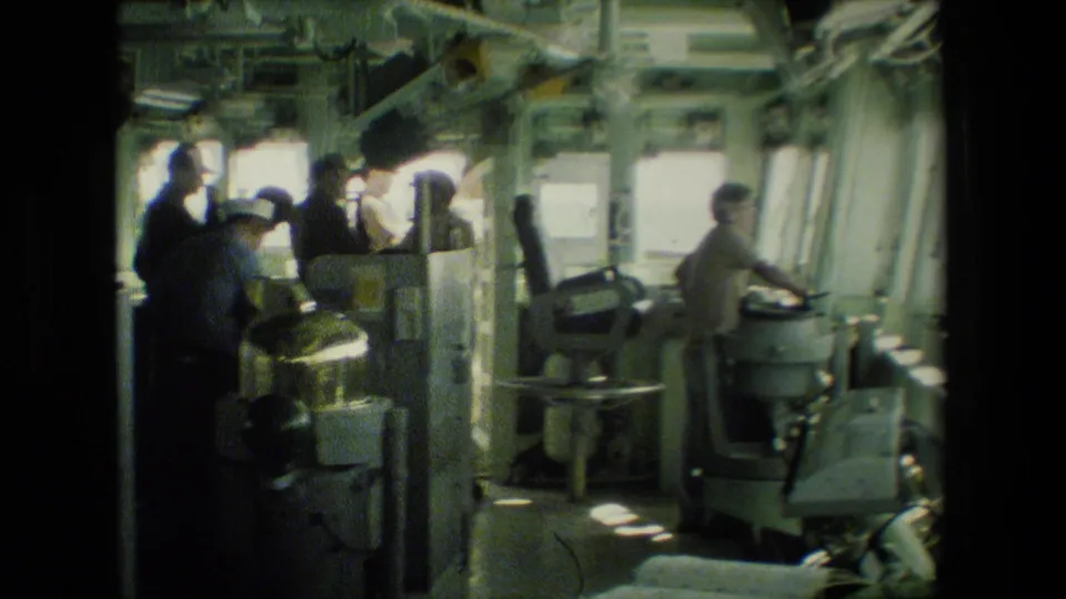 Uniformed sailors appear inside a naval ship workspace with consoles, seats, windows, and overhead equipment visible.