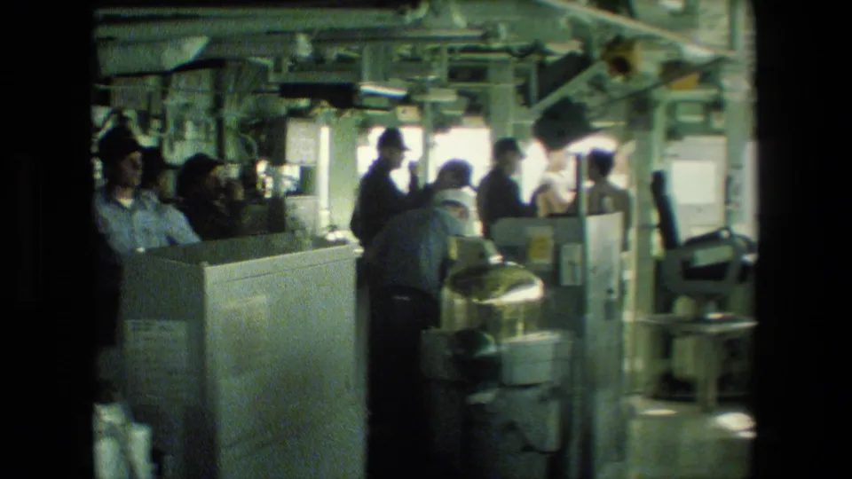 Several sailors work or observe near shipboard consoles and equipment in a compact naval interior with bright window light behind them.