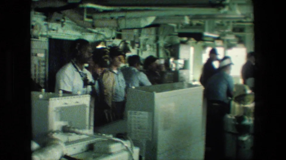 A group of uniformed sailors stands inside a naval ship interior near consoles, monitors, windows, and overhead machinery.