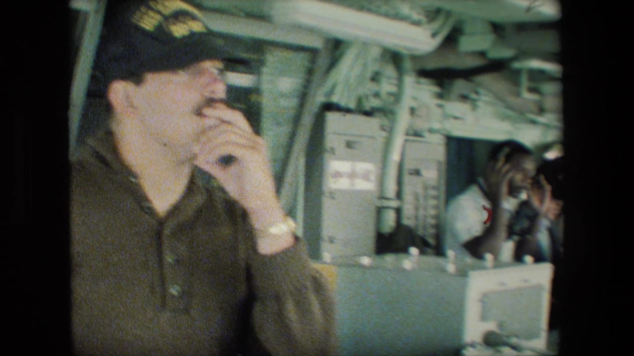 A man wearing a dark naval-style cap speaks into a handset or microphone near control panels and crew members inside the submarine control-room setting.