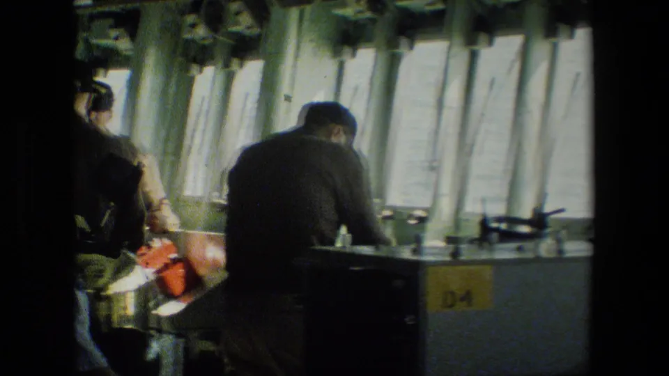 Crew members are seen from behind and partly in shadow as they work near a console and vertical panels in the submarine control-room area.