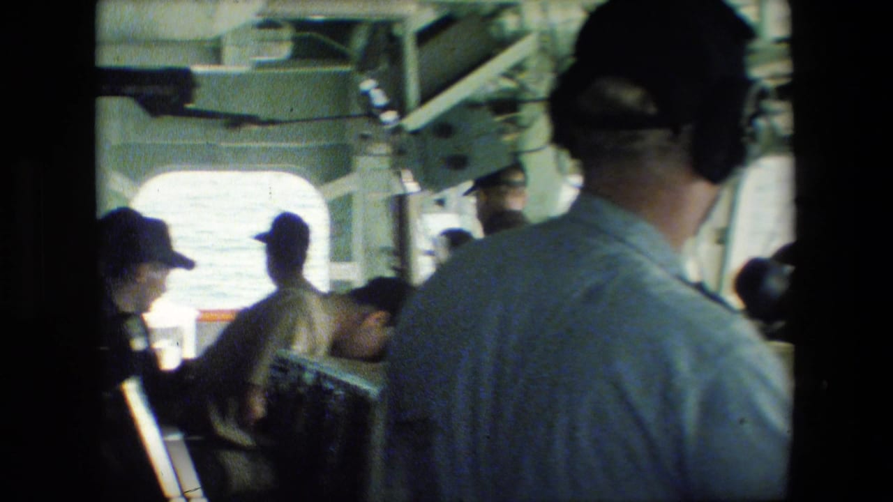 Several men in caps stand and work near control stations inside a bright-windowed vessel interior.