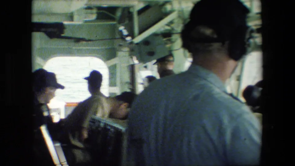 Crew members appear at control stations inside a naval vessel interior, with a man seen from behind in headphones and others silhouetted near windows.