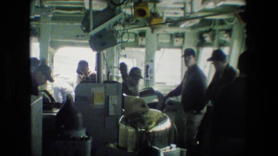 The 8mm frame shows men in caps moving and working around equipment inside a vessel interior, with daylight through rectangular windows.