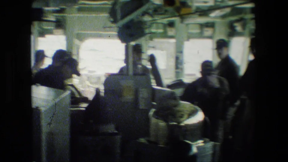 A dim shipboard interior shows men seated and standing around equipment, with bright exterior light through windows and metal structure overhead.